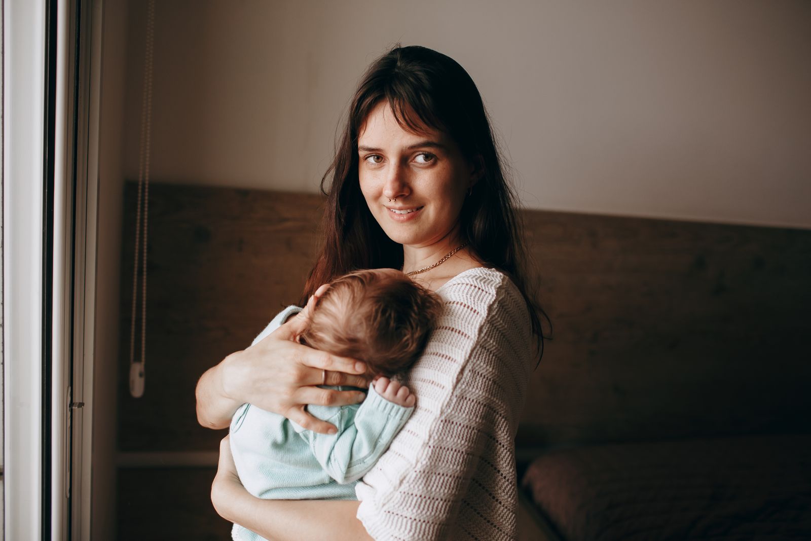 A mother holding her sleeping baby beside a window at home